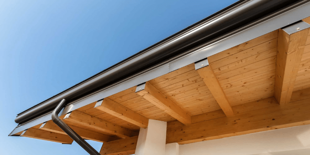 Low-angle view of a black round gutter and downspout mounted beneath a cedar soffit against a blue sky in Central PA
