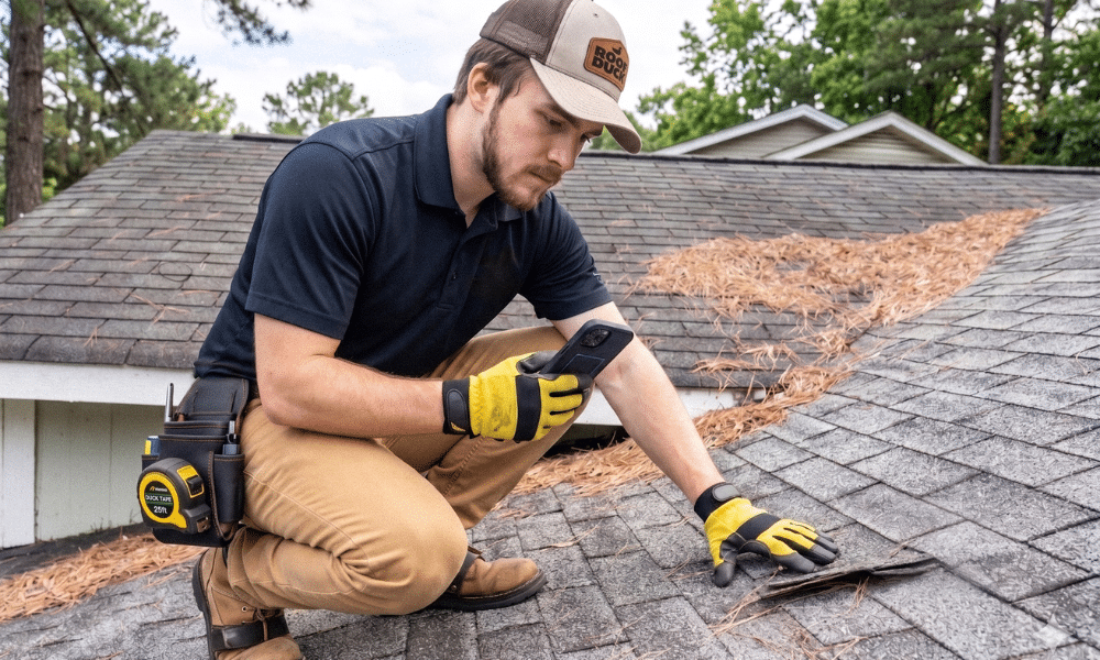 Roof Duck inspector in a branded cap kneeling on an asphalt shingle roof, lifting a damaged shingle with yellow work gloves