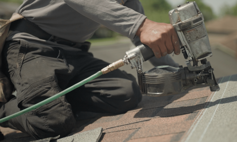 Roof Duck Worker holding a nail gun close to the surface while nailing down charcoal asphalt shingles during a roof repair in Central PA